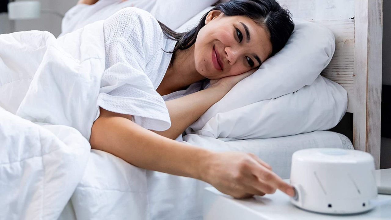Person in bed under covers pressing a button on a white noise machine.