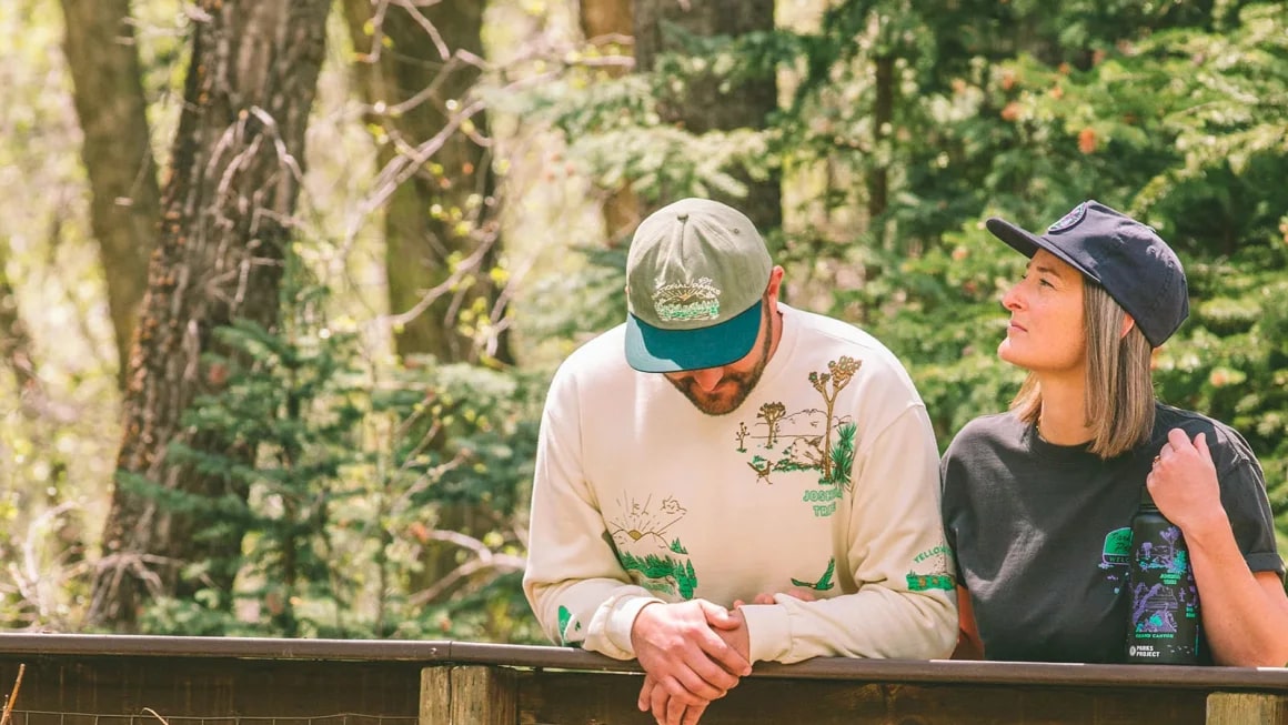 Two people standing on a bridge in nature wearing Parks Project shirts and hats.