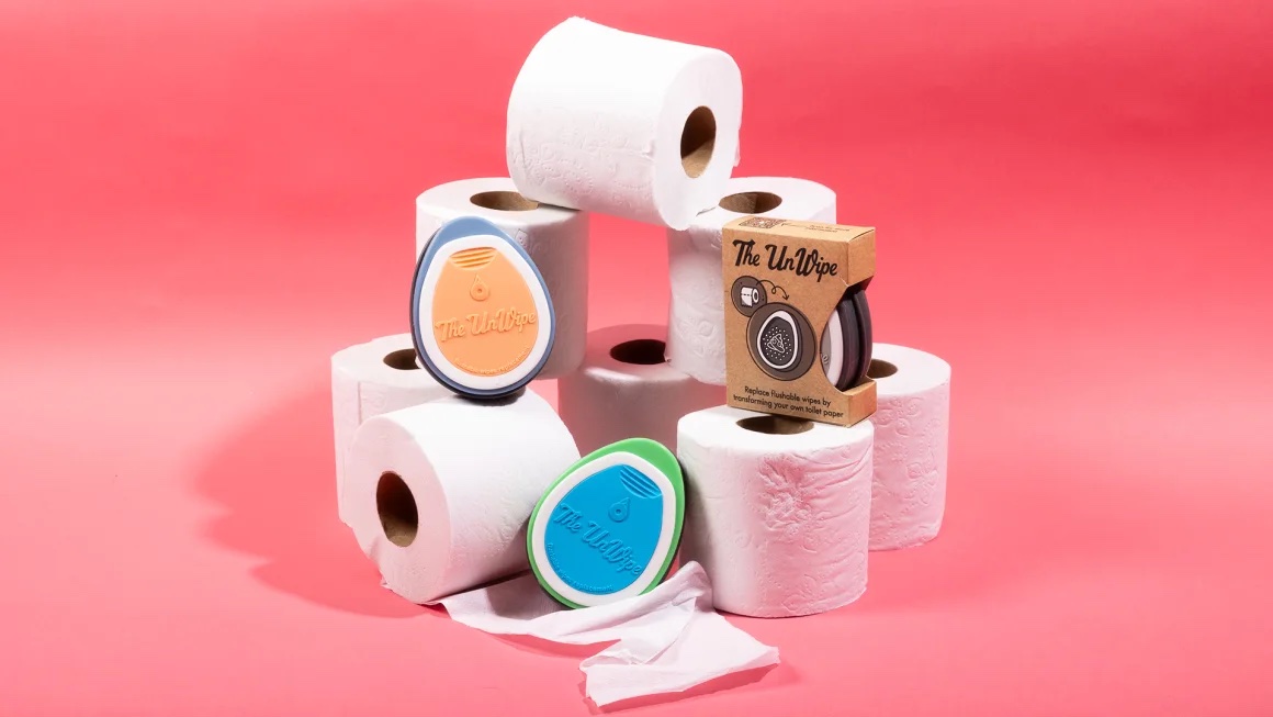 The UnWipe tool nestled on top of a semi-pyramid of toilet paper against a salmon-pink backdrop